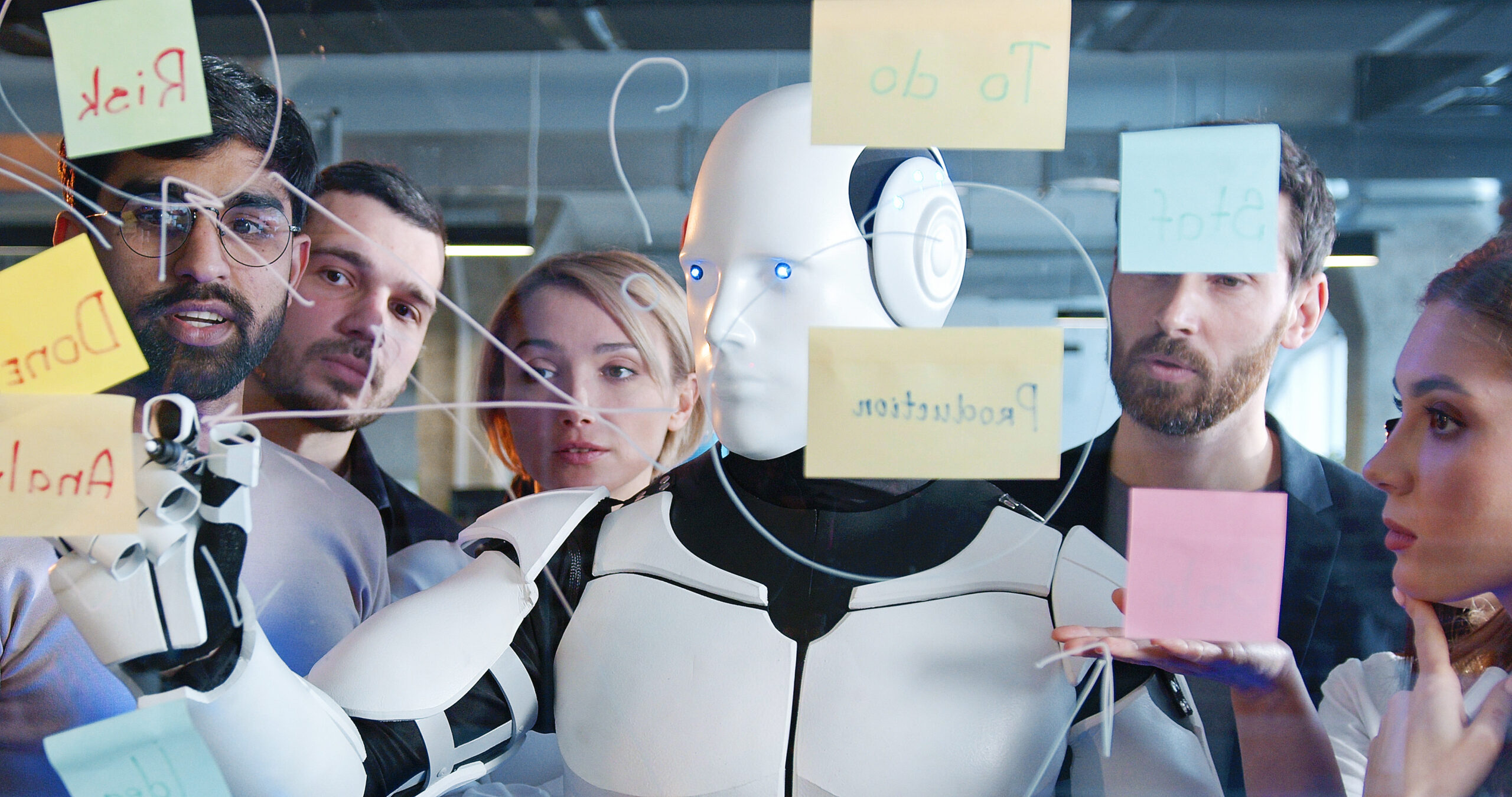 Diverse team of project discussing plans and to-do notes with robot. Teambuilding of multiethnic colleagues. Coworkers brainstorming together with humanoid. Management strategy. Digital transformation