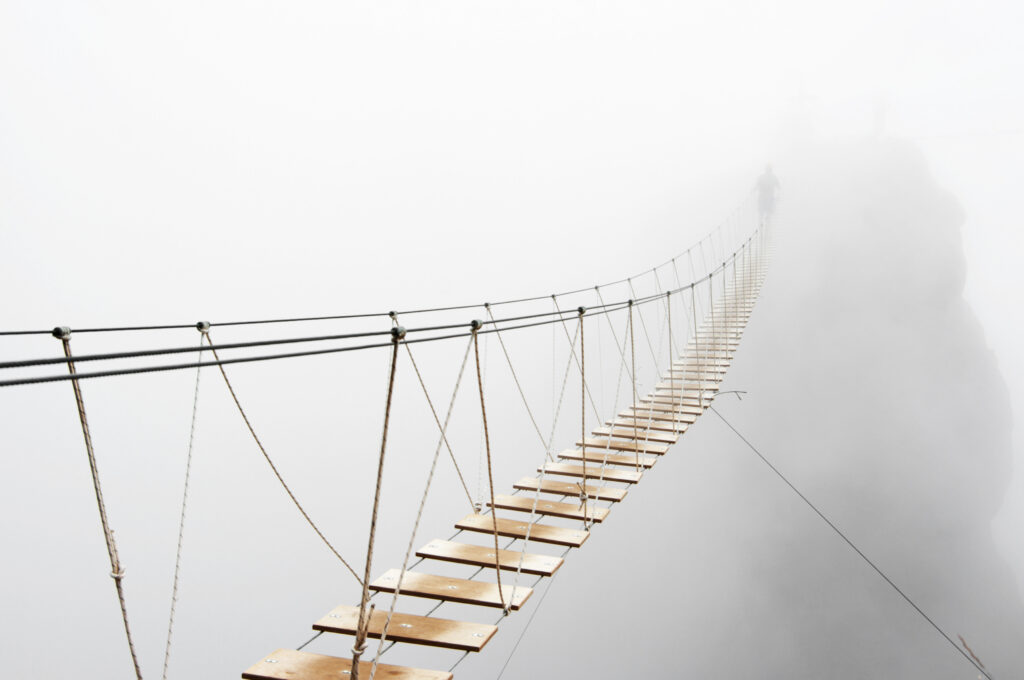 A suspension bridge made of ropes and wooden planks disappears into a dense fog, representing uncertainty.