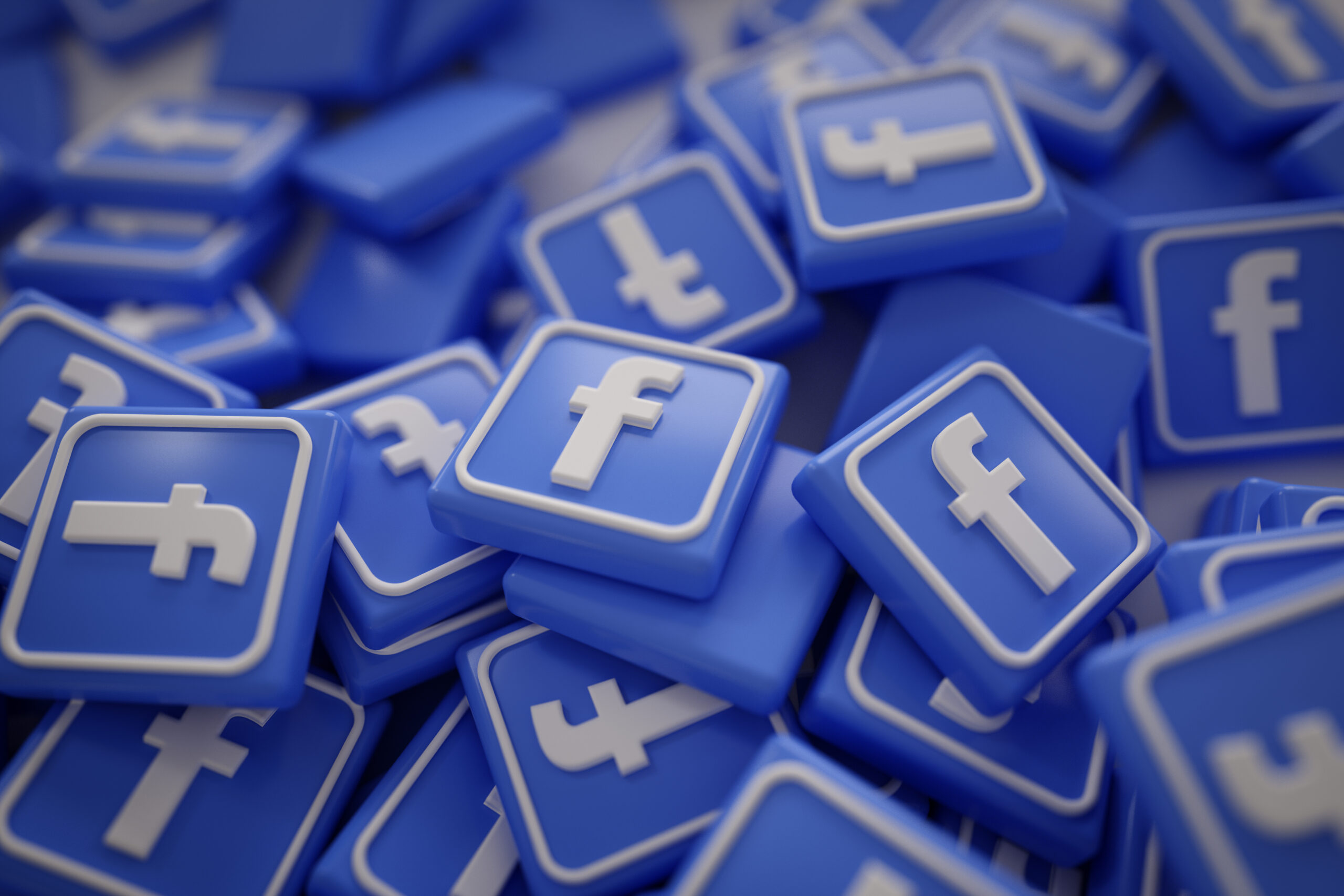 The image shows a collection of three-dimensional Facebook "f" logo icons in blue and white. The icons are scattered and piled up, with some facing upwards and others at various angles, creating a sense that there are numerous such icons in the image. The "f" is the well-known logo of Facebook, a popular social media platform. The colors are consistent with the Facebook brand's color scheme.