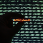 A hand is silhouetted against a screen displaying binary code with the word "RANSOMWARE" highlighted in red.