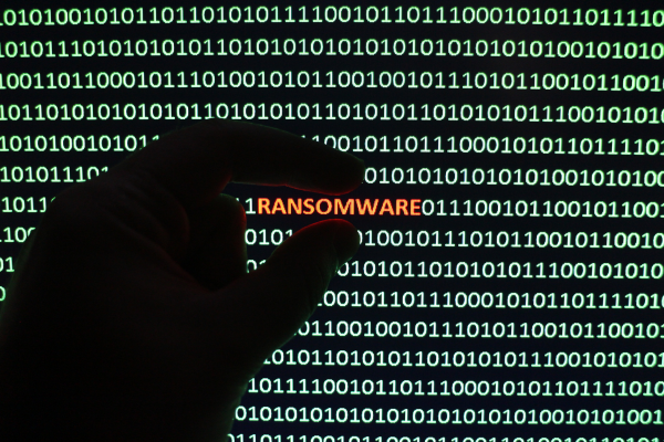 A hand is silhouetted against a screen displaying binary code with the word "RANSOMWARE" highlighted in red.