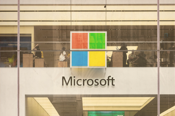 The image shows the facade of a modern Microsoft store, with large glass windows revealing a busy interior. The iconic Microsoft logo in bright colors is visible above the entrance, symbolizing the tech giant's branding.