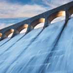 The image depicts a large dam with arch structures, where cascading water flows down its spillway.