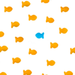 Numerous orange goldfish-shaped crackers are scattered across a white background with a single blue goldfish cracker standing out in the center, creating a visual metaphor for uniqueness.