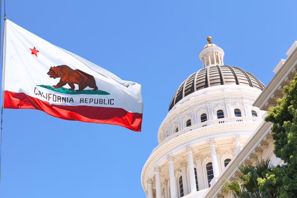 The image shows the California state flag. In the background, the domed white structure of the California State Capitol in Sacramento can be seen.
