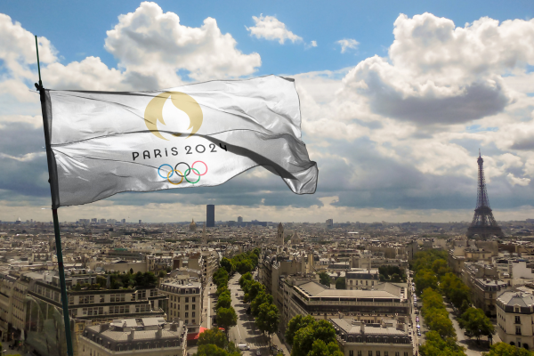 A photo of the Paris skyline on a day with scattered clouds, featuring the iconic Eiffel Tower in the distance. Prominently displayed in the foreground is a flag with the 'Paris 2024' Olympic Games logo, fluttering in the wind.