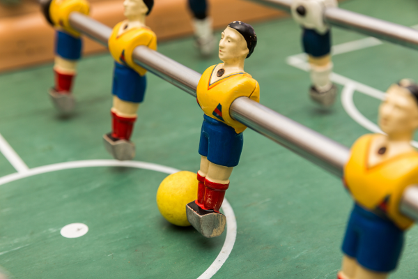 The image shows a close-up view of a foosball table, featuring a yellow foosball in play. One of the miniature players is dressed in a blue uniform with red socks and red, strapped shin guards.