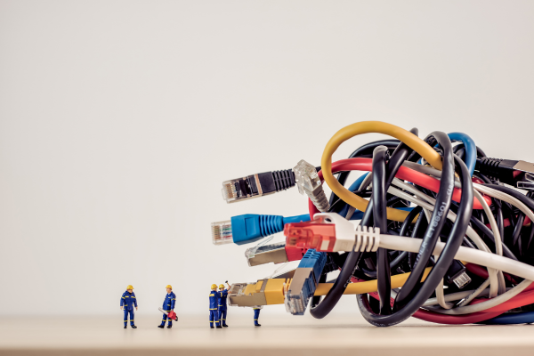 A group of miniature construction workers stands in front of a large, tangled bundle of various computer cables, including Ethernet and USB cords, appearing as if they are preparing to untangle and organize them.