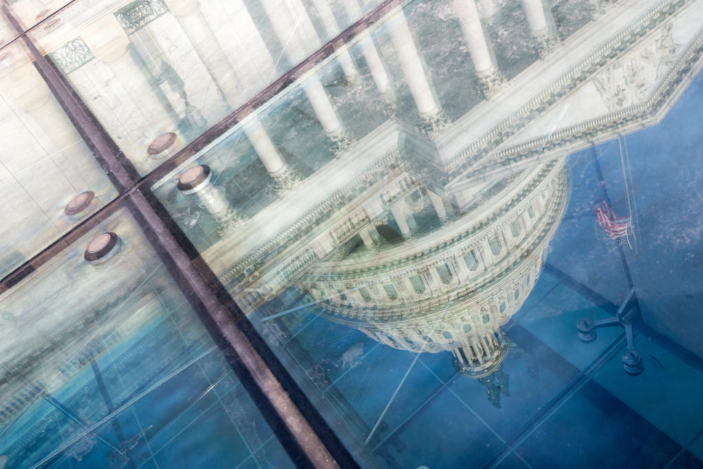 Reflection of the U.S. Capitol dome and columns in angled glass panels, with an American flag visible against the blue sky.