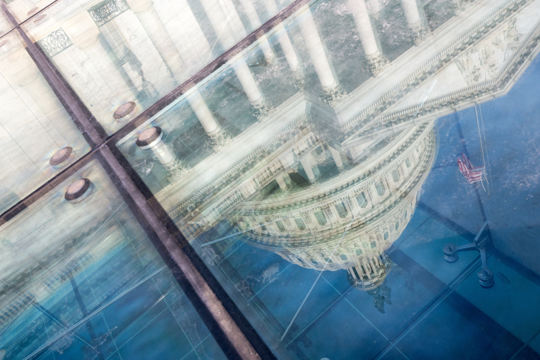 Reflection of the U.S. Capitol dome and columns in angled glass panels, with an American flag visible against the blue sky.